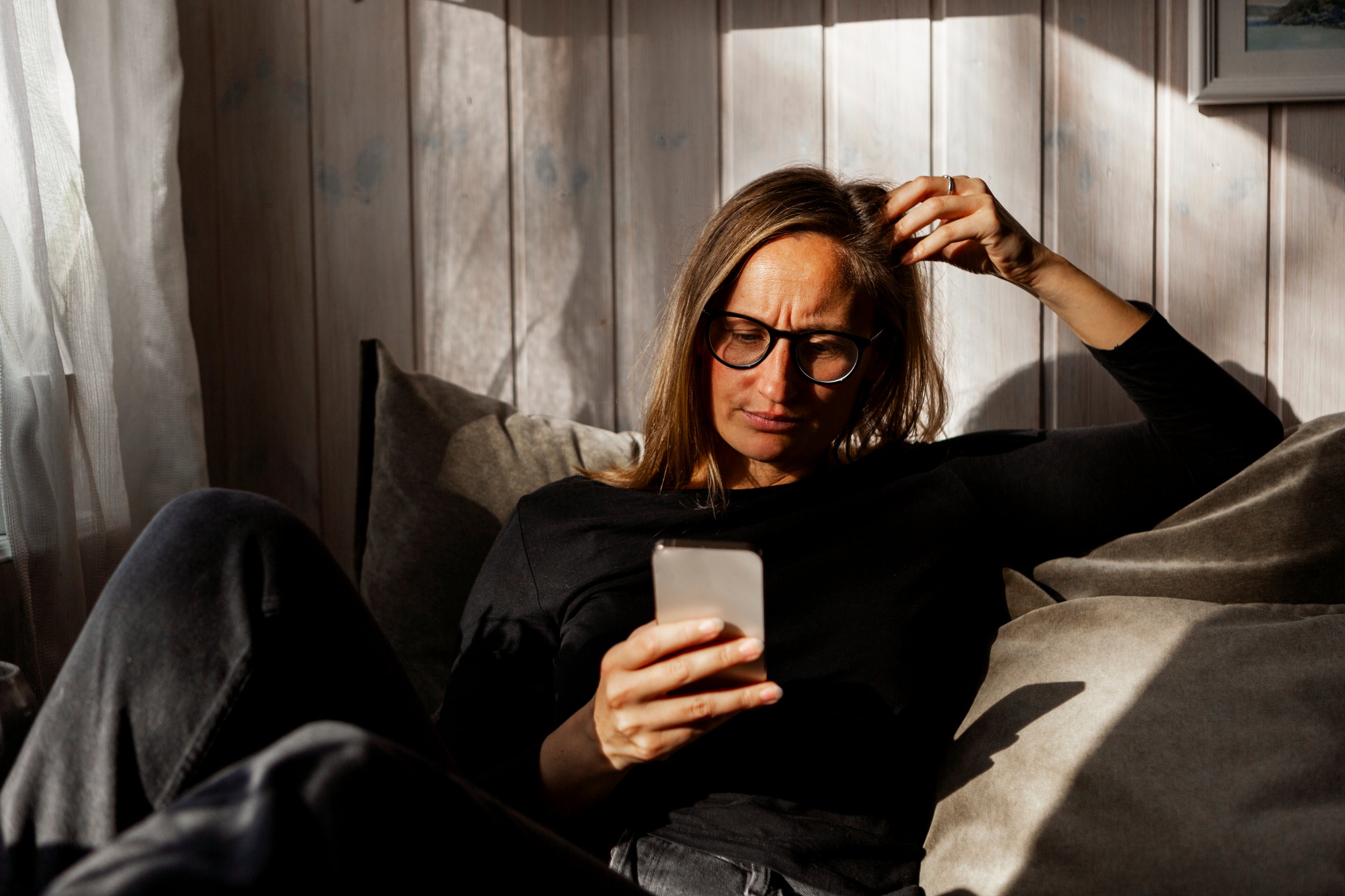 Woman with long hair and glasses sitting in a sofa while looking down at a mobile phone
