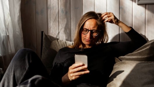 Woman with long hair and glasses sitting in a sofa while looking down at a mobile phone